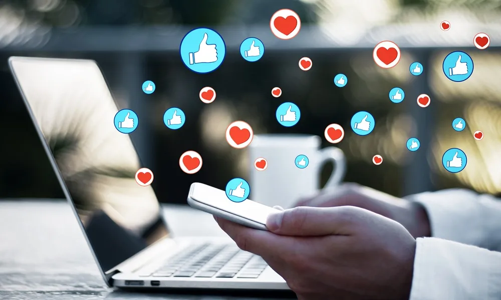 Social commerce: como transformar seguidores em compradores nas redes sociais? Mãos segurando celular em frente a um notebook com ícones de curtidas e corações flutuando.